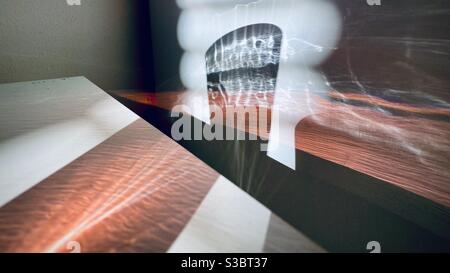 Patterns of light created by sunlight passing through a window blinds and glass of red liquid, refracting across white table and wall to create abstract background Stock Photo