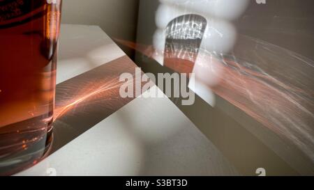 Patterns of light created by sunlight coming through a window blinds and through a glass of red liquid, refracting across white table and wall to create abstract background Stock Photo