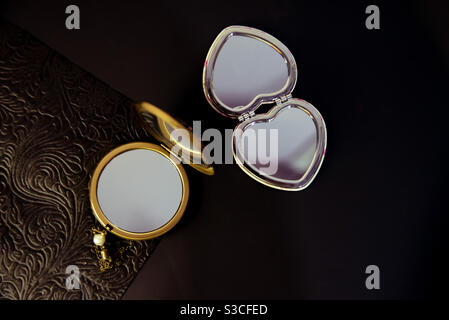 a contrast between a gold round pocket mirror and a silver heart-shaped pocket mirror on a half-textured black background Stock Photo