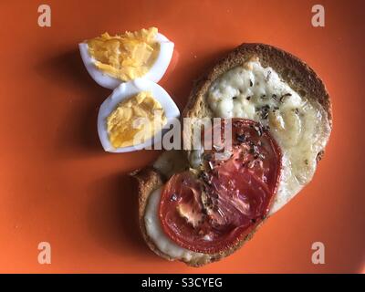 Slice of toasted bread with melted cheese on it and cooked slice of tomato served with herbs and a boiled egg next to it Stock Photo