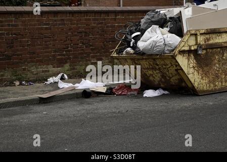 A household skip left on the roadside that is over flowing with rubbish garbage and litter all over the road with copy space Stock Photo