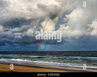 Beautiful rainbow over the sandy beach, standing out against a grey stormy sky leading into the sea, waves, nature, Australia Stock Photo