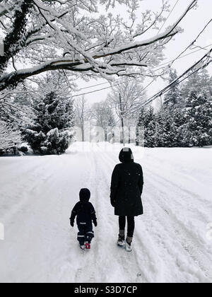 A mother and three year old child walking down a snowy street together during a snowfall in New Jersey.