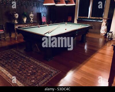 A classic picture of the billiard room a pool table in a dark wood room. Two pool cues on the table along with colored balls ready for a game Stock Photo