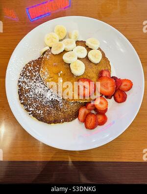 And inviting plate of powdered sugar covered pancakes with sliced bananas and strawberries on a white plate set at a tan-colored table reflecting backwards a red neon sign saying breakfast Stock Photo