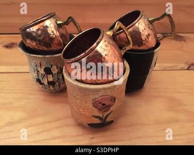 Three copper cups sitting in ornate clay pots on a wood plank, wooden slats in the background. An elegant arrangement of two trios, some functional, others decorative. Canada Stock Photo