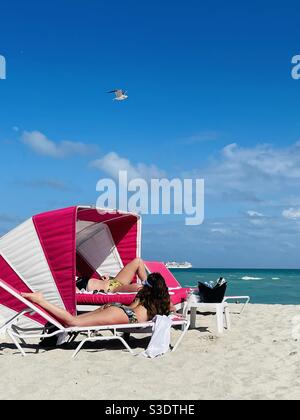 Women sunbathing under a pink and white cabana umbrella with cocktails on the sandy beach looking Toward a cruise ship on the horizon of the turquoise water In Miami South Beach, Florida, USA Stock Photo