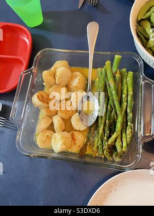 Home cooked scallops and asparagus in a garlicky butter sauce in a glass serving dish With a spoon set on a dark blue tablecloth Stock Photo