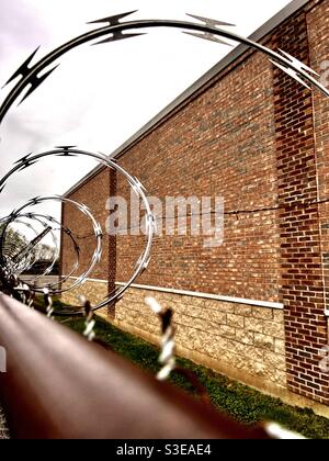 Looking through the barbed wire to the other side; view of a gloomy brick building through cruel looking barbed wire atop a metal fence. Stock Photo