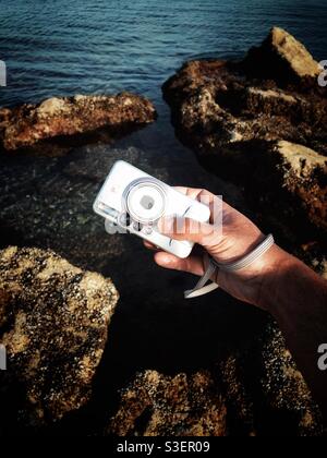 Man holding a point and shoot camera over the rocky shore Stock Photo