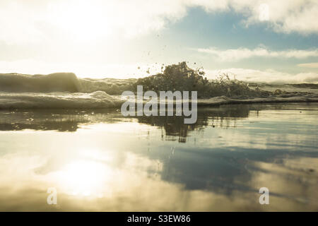 Low angle shot of a wave washing up onto beach as the late afternoon sunny sky reflects on the wet sand. Stock Photo
