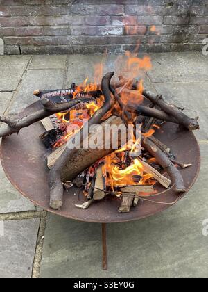 Wood burning in rusty fire pit on patio stones by brick wall Stock Photo