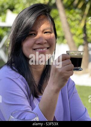 Filipino Asian woman holding and drinking a cup of espresso coffee in a backyard during a sunny summer day Stock Photo