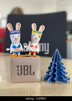 Two toy Raving Rabbits standing on top of a golden JBL Go Bluetooth speaker, with a green paper Christmas tree on an office desk Stock Photo