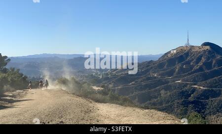 Horse riders kicking up dust on trail through Griffith Park with Hollywood sign visible on the right Stock Photo