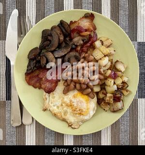 Fried breakfast with egg, bacon, fried potatoes, mushrooms and baked beans on a green plate with stainless steel cutlery at side Stock Photo