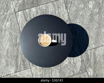 Overhead view of a cup of coffee on the plain dark top of a garden table standing on the grey porcelain paving slabs of patio Stock Photo