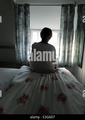 A lone woman man sitting alone in her bedroom and looking out of a window in a depression, anxiety or loneliness concept Stock Photo