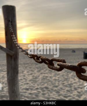 A rusty chain is attached to a wooden pole on the beach, in front of the sunset in the background Stock Photo