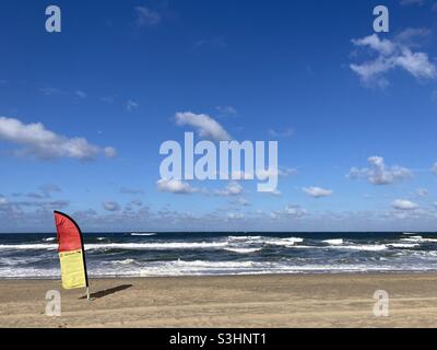 A red yellow flag stands on the beach by the sea in a strong wind in sunny weather and blue skies Stock Photo