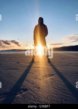 A person stands on the beach with the sun setting between their open legs in Scotland Stock Photo
