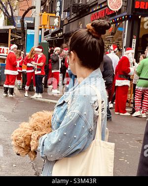 A woman holding a cute fluffy dog watches people dressed as Santa Claus at an intersection in New York City during SantaCon NYC December 2021 Stock Photo