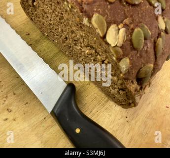 Half a loaf of brown bread studded with pumpkin seeds, together with a serrated bread knife Stock Photo