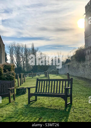 The low winter sun sits behind a stone building, setting shadows over the graveyard. A contemplation bench sits in the foreground. No people Stock Photo