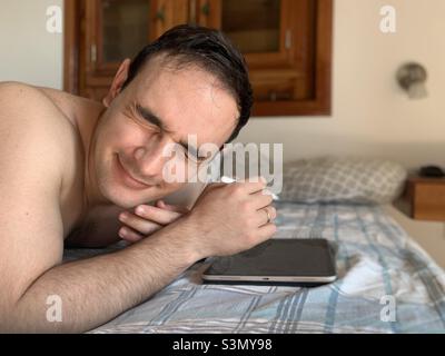 Photophobia. Young adult man working on tablet while laying still in bed closes his eyes because of the sunlight that entered when a door was opened. Stock Photo