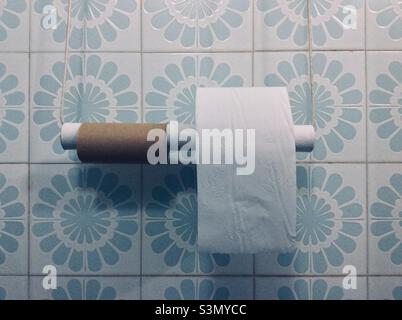 Toilet paper rolls, empty and new, on DIY holder in a bath, backdropped by light-blue tiles wall. Soft filtered, vintage, moody filmic style Stock Photo