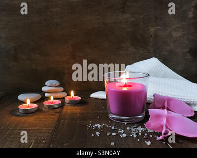 There are lit aromatic candles on a wooden table, sea salt for a relaxing bath is scattered from a pink bag, a pebble stone pyramid is in the background, copy space. Stock Photo
