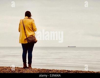 Woman in a bright mustard yellow coloured coat facing out to sea Stock Photo