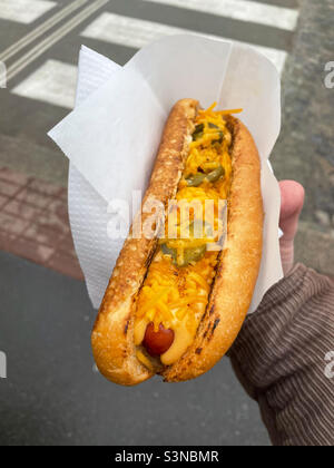 Male hand holding hot dog with jalapenos and grilled cheddar in a paper napkin outdoors on the street. Stock Photo