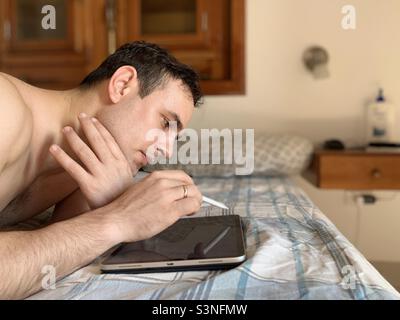 Designer working on iPad lying on bed with early morning sunlight. Creative work. Stock Photo
