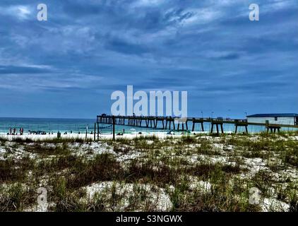 A view of sand dunes covered in beach grass and a white sand beach in the distance and a fishing pier stretching out over the ocean / gulf / sea, landscape, summer beach scene Stock Photo