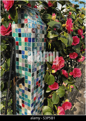 Brightly coloured mosaic gatepost surrounded by pink roses Stock Photo