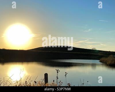 The sun beginning to set behind a hill. The reflection of the sun can be seen in the lake at the forefront. Stock Photo