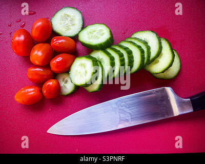 Sliced cucumber with baby plum tomatoes wit a knife on a red chopping board. Stock Photo