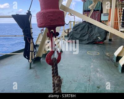 Close view on red hook from bunker crane secured by rope to green deck on the merchant container vessel. In background is calm sea, blue sky and stowed containers. Stock Photo
