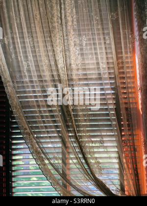 Lace curtains hanging over partially closed blinds with light filtering in. Stock Photo