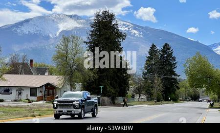 HAMILTON, MT, MAY 2022: black pick-up truck driving on rural road, with trees behind, and small white clouds over mountains in background Stock Photo