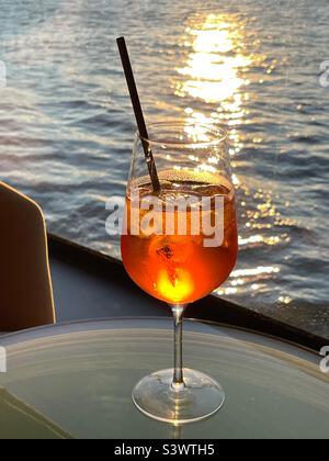 Glass of Aperol Spritz against a background of the setting sun over water Stock Photo