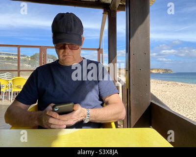 Remote working, man sitting in a beach cafe working on his mobile phone Stock Photo