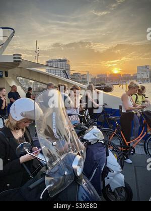 Amsterdam commuters on bicycles and mopeds leave the city centre on the free evening ferry and scroll their devices during a glorious sunset Stock Photo