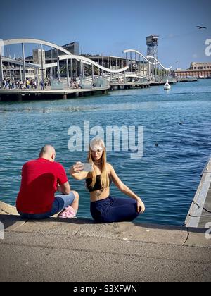 A woman takes a selfie sitting on the waterfront harbour wall overlooking the popular tourist attraction of Port Vell in Barcelona Stock Photo