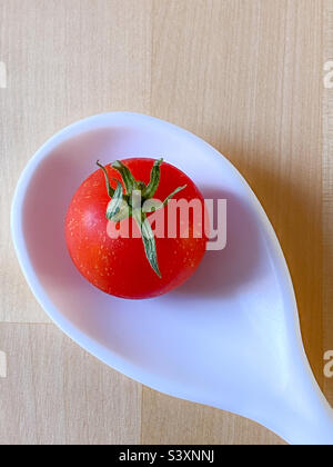 Still life image of a single cherry tomato in the bowl of a white spoon on a wooden table Stock Photo