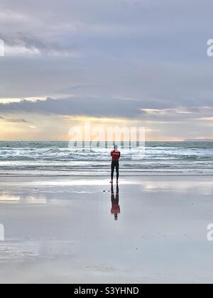 The lifeguard and his reflection in the wet sand at sunset on a warm summers evening on Saunton Sands, Devon, England. The fit young man is a coach and lifeguard watching for swimmers Stock Photo