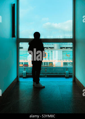 A boy stood looking out of the window of a tall building at the end of a corridor Stock Photo