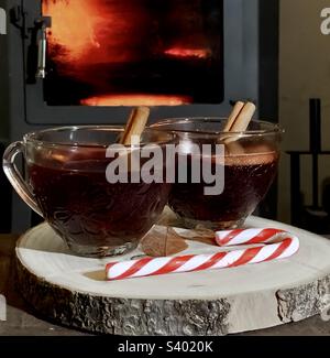 2 punch glasses full of mulled wine on a wood platter with a candy cane, in front of a roaring wood burner. Stock Photo