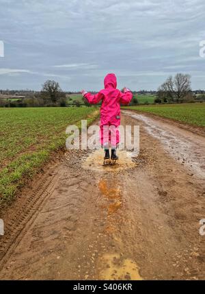 Rear view of girl jumping in muddy puddle on rural road in the rain. Stock Photo